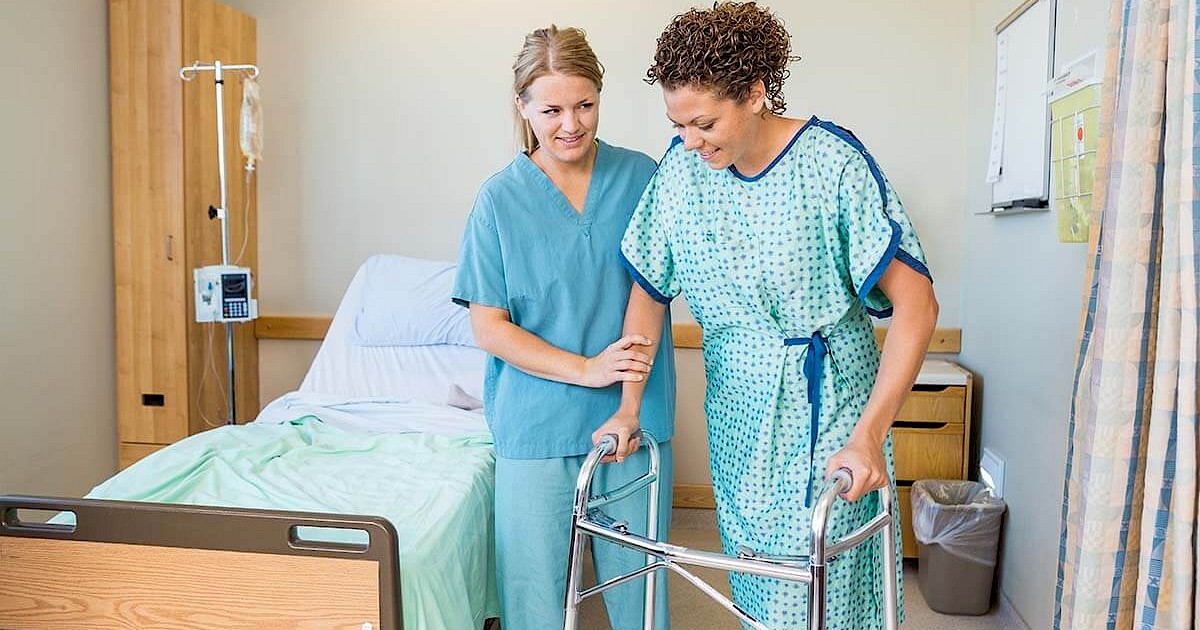 Nurse assisting patient in hospital gown to walk with walker